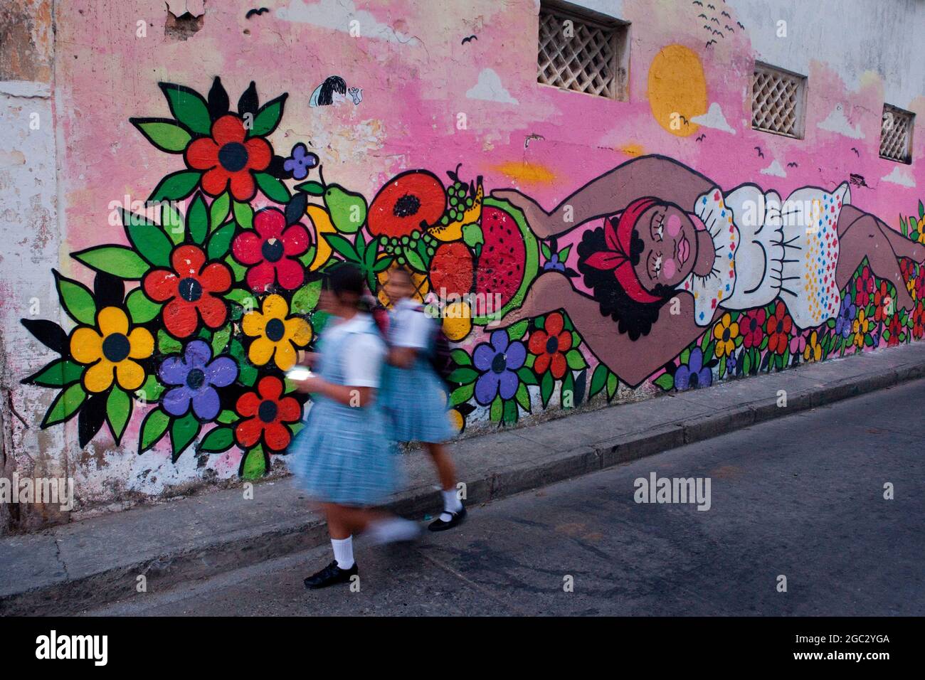 Local school kids stroll past a colourful street art mural of a palenquera fruit vendor in the Getsemani neighbourhood of Cartagena, Colombia. Stock Photo