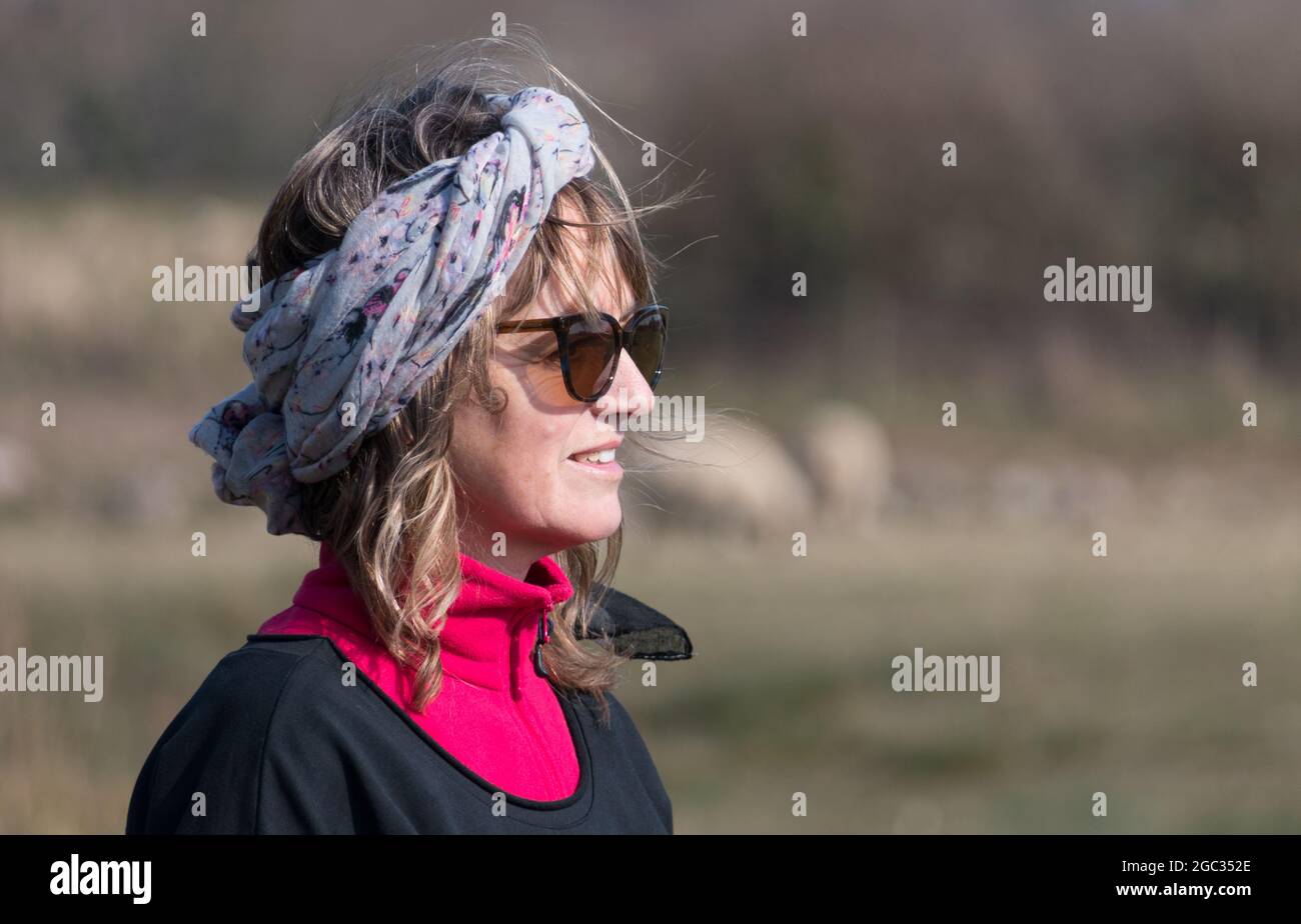 An attractive lady looks right with a headscarf long hair on windy but sunny day wearing sunglasses.She smiles happily Stock Photo