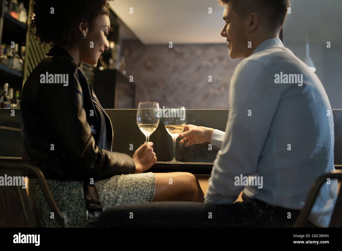 Romantic interracial couple drinking white wine sitting at the pub restaurant counter. Main focus on the wineglasses. Girlfriend and boyfriend clinkin Stock Photo