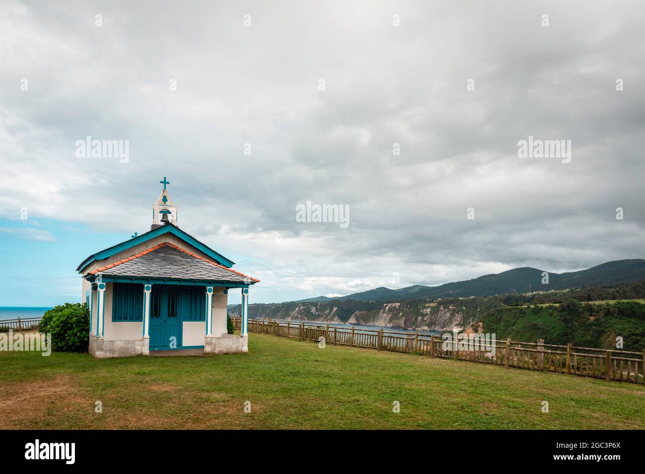 In the foreground a white and blue chapel in a meadow, in the background the cliffs of the Spanish coast and the Cantabrian Sea. Stock Photo