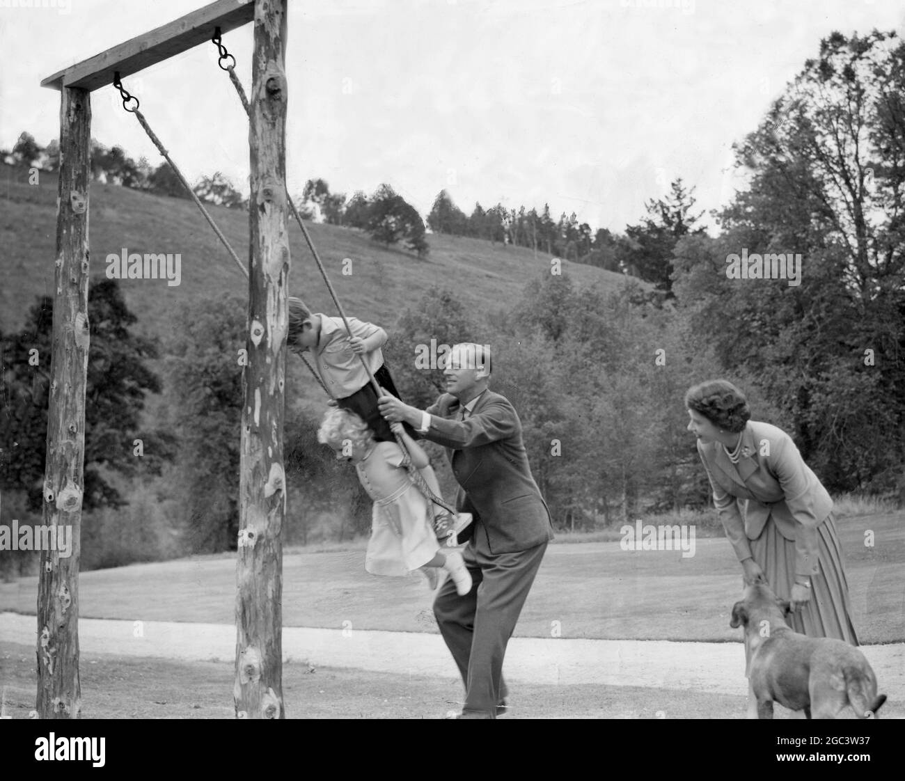 The Royal Family at Balmoral, Scotland - June 1956 Prince Philip, The Duke of Edinburgh pushes his children, Princess Anne and Prince Charles, on a swing, with Queen Elizabeth looking leaning down to a pet dog. Stock Photo
