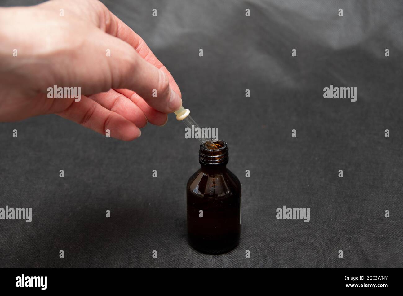 A hand holds a glass dropper with a yellow rubber tip with liquid over a dark brown bottle on a dark trendy background close-up. Stock Photo