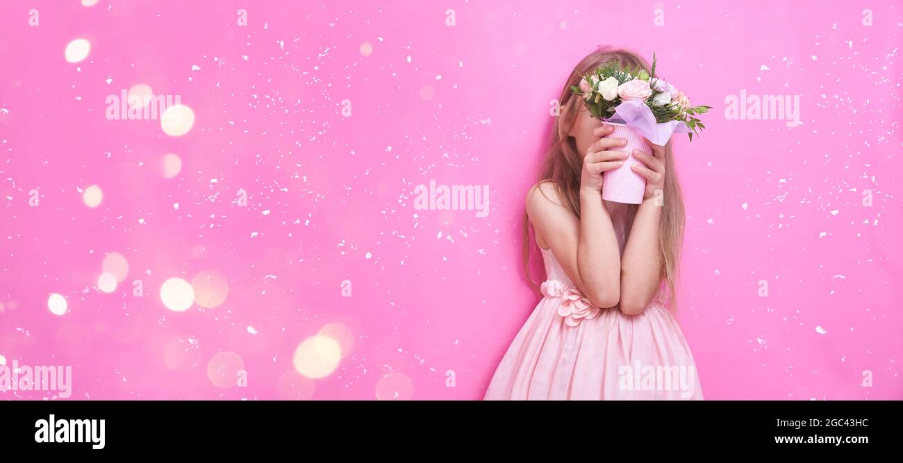 A little girl in pink dress with bouquet of flowers in a paper cup, hid behind the flowers. The girl is shy and sniffs the flowers on pink banner. Stock Photo