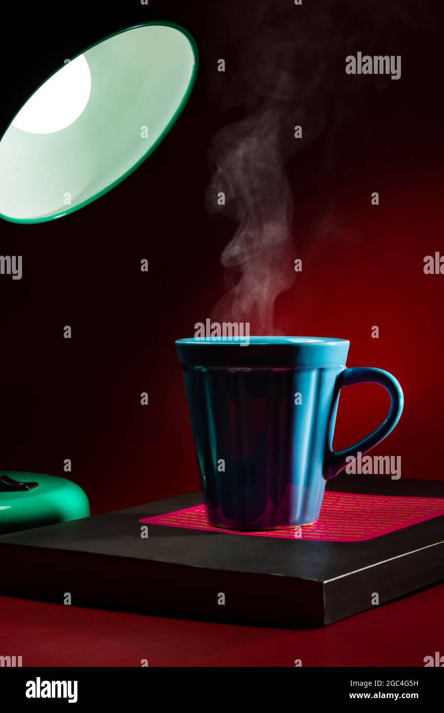A lamp illuminating a blue mug with coffee or tea, over a closed book and a red background. Stock Photo