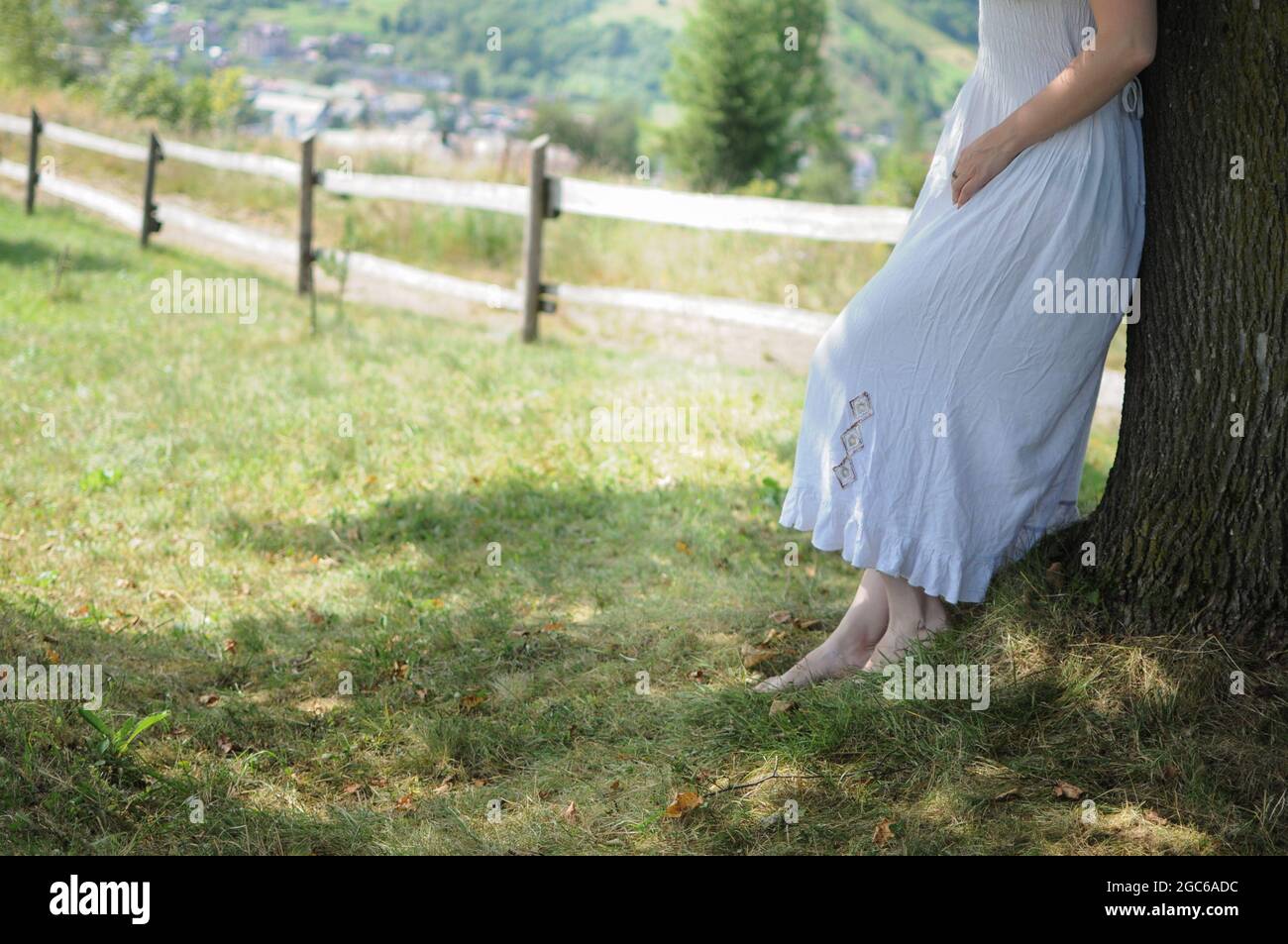 Young woman in white dress is standing near a tree in meadow. Copy space ethnic Stock Photo