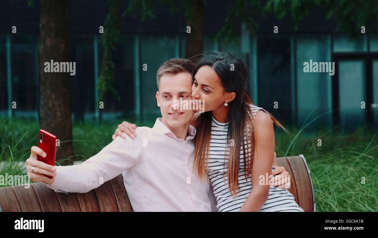 Romantic multiethnic couple sitting on bench in park and taking selfie. They smiling to the camera. Stock Photo