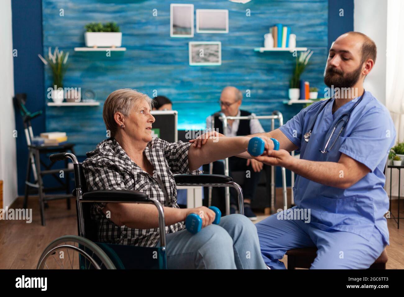 Male nurse helping senior retired disabled woman in wheelchair to rehabilitate using dumbbels during recovery session in nursing home. Elderly woman exercising physiotherapy with social worker Stock Photo