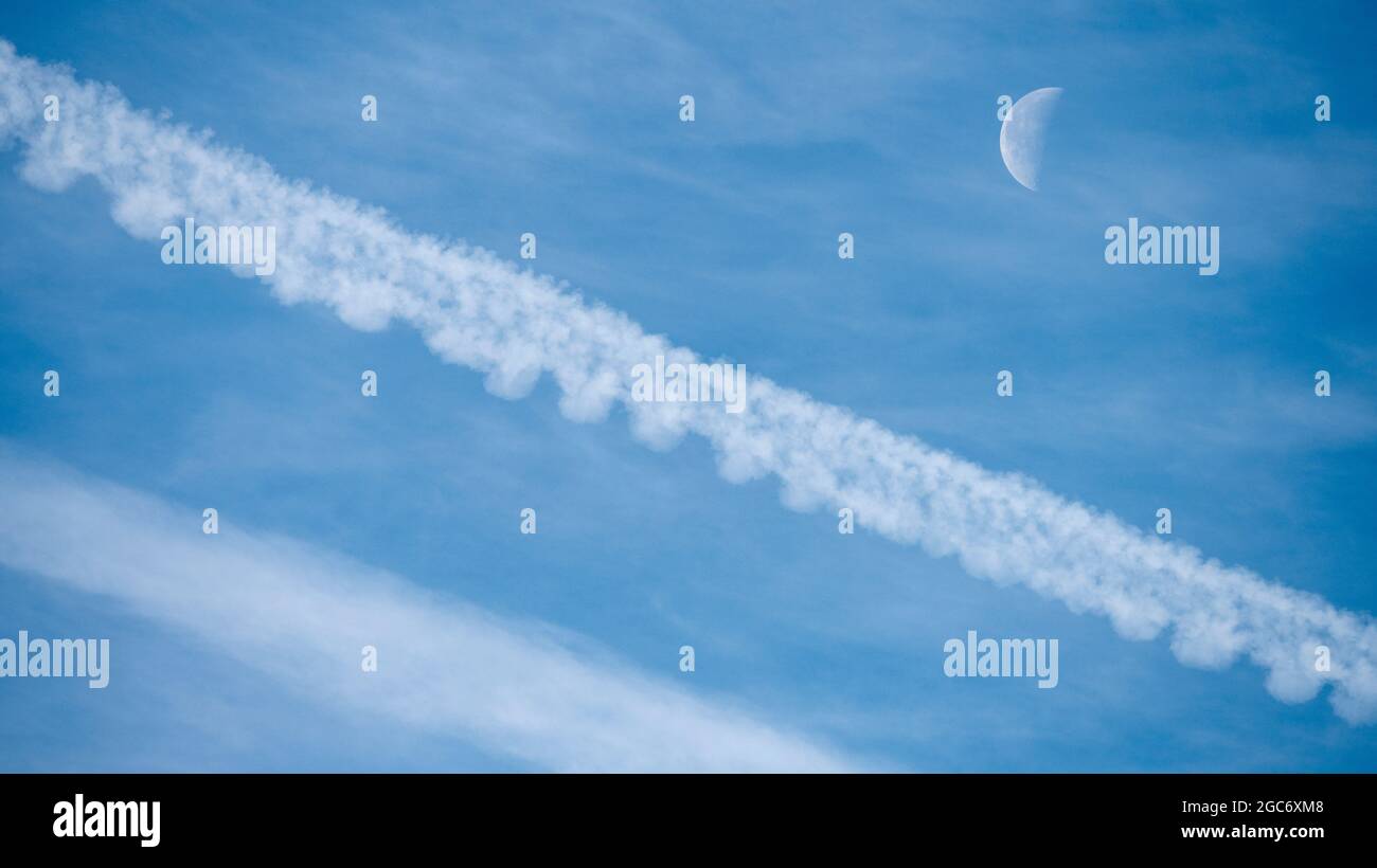Airplane contrails and waning moon against blue sky Stock Photo