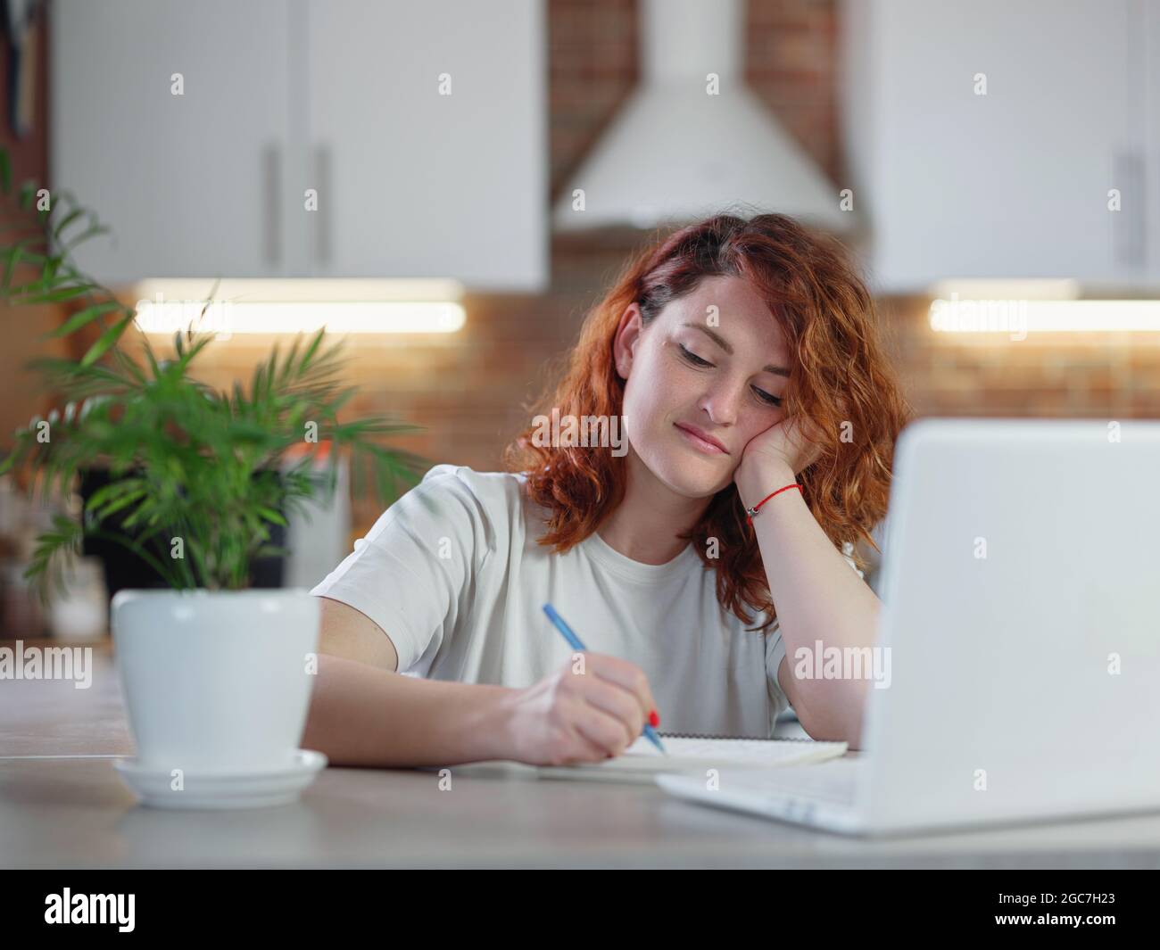 A beautiful young redheaded woman sits in the kitchen at the table with ...