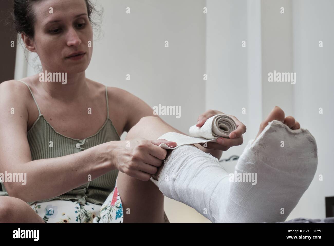 Woman sitting on a bed puts an elastic bandage on a broken leg. The leg is in a plaster cast. home rehabilitation after a broken leg. High quality pho Stock Photo
