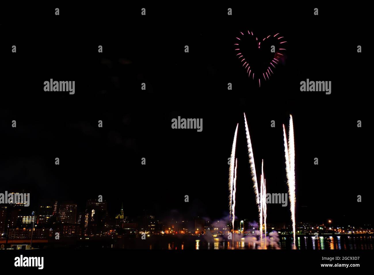 Heart shaped fireworks sideways over a city skyline at night. Other fireworks rise towards the heart. Stock Photo