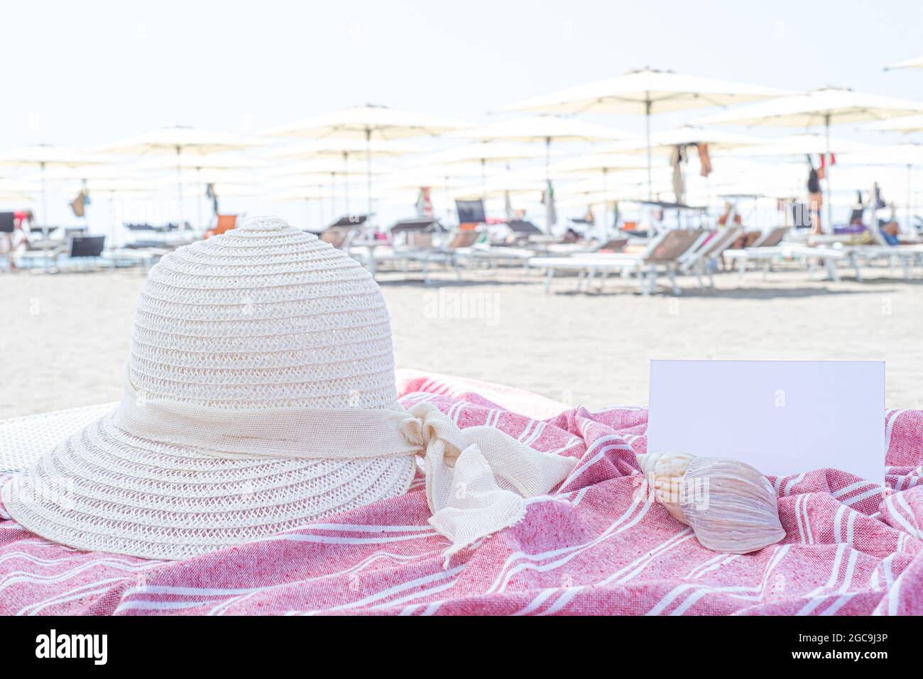 Summer vacation still life. White straw hat, seashell on red and white towel.Beach umbrellas in the background. Blank invitation card or greeting card Stock Photo