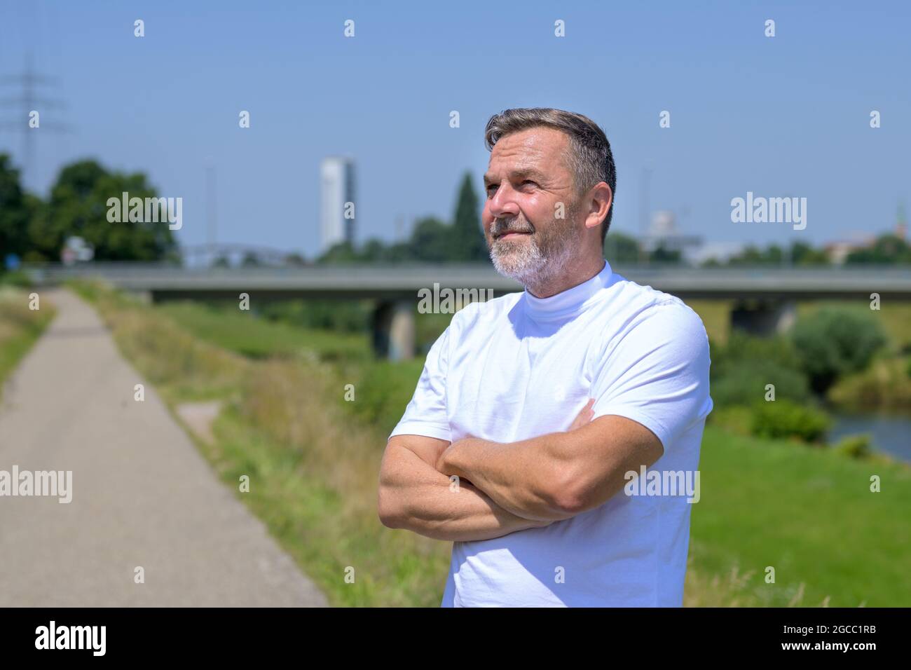 Thoughtful elderly man standing with folded arms staring up into a sunny sky on a rural footpath alongside a river in an upper body view Stock Photo