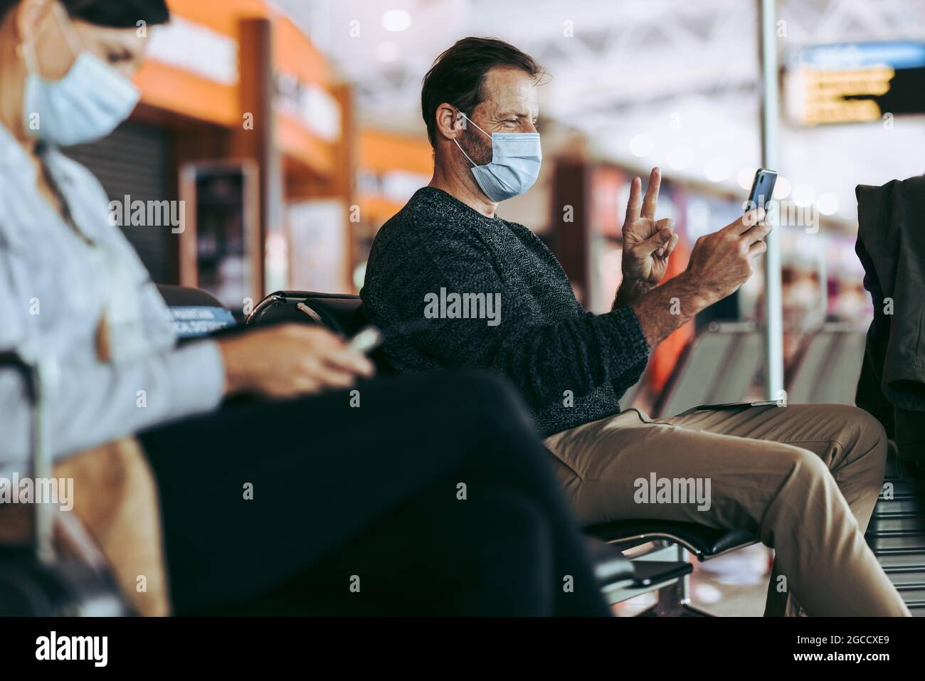 traveler during pandemic video calling on mobile phone while waiting at airport. Man in face mask sitting at airport terminal and video calling. Stock Photo