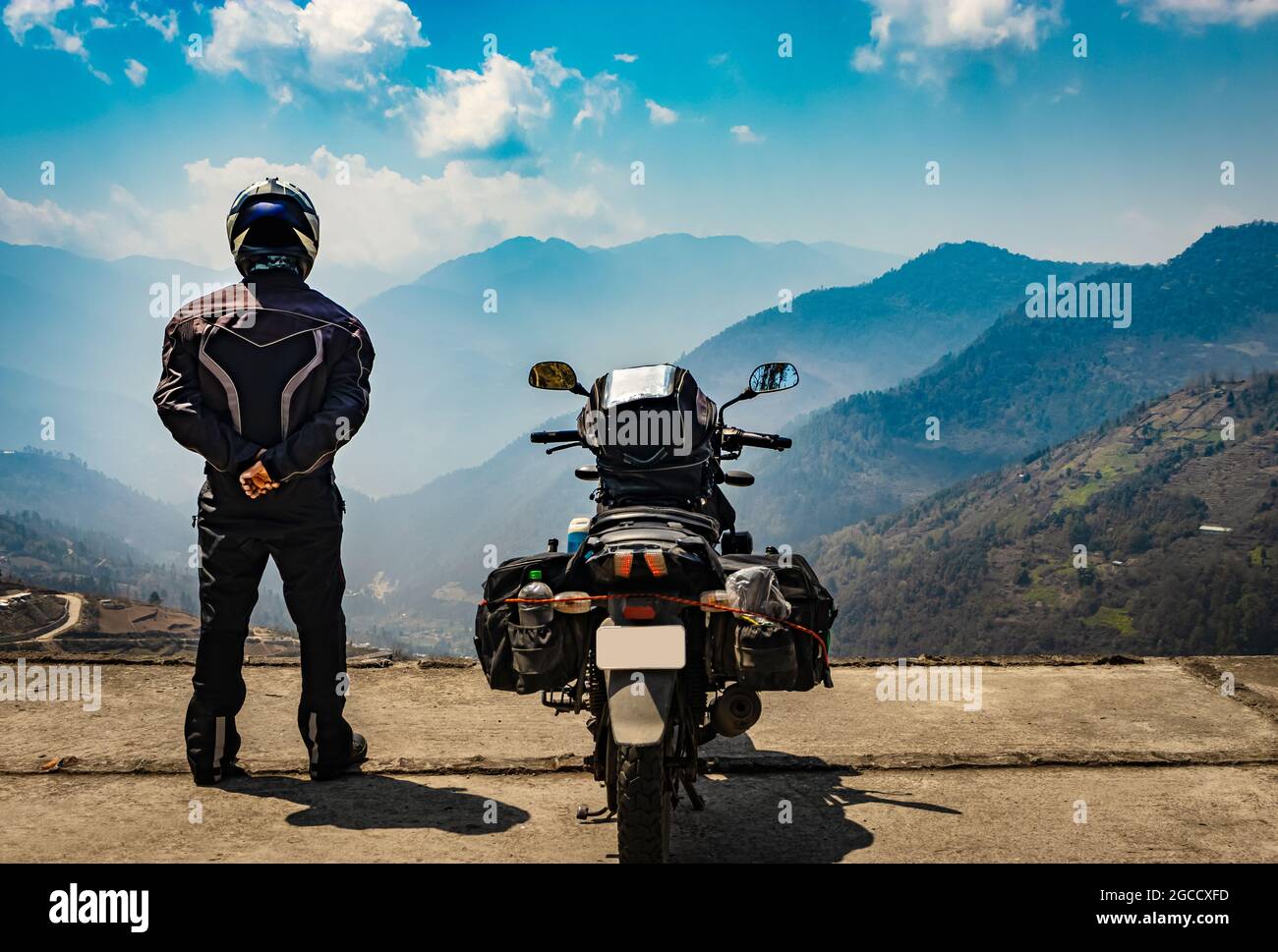 man motorcyclist watching valley from hill top with his loaded motorcycle and pristine natural view image is taken at bomdila arunachal pradesh india. Stock Photo