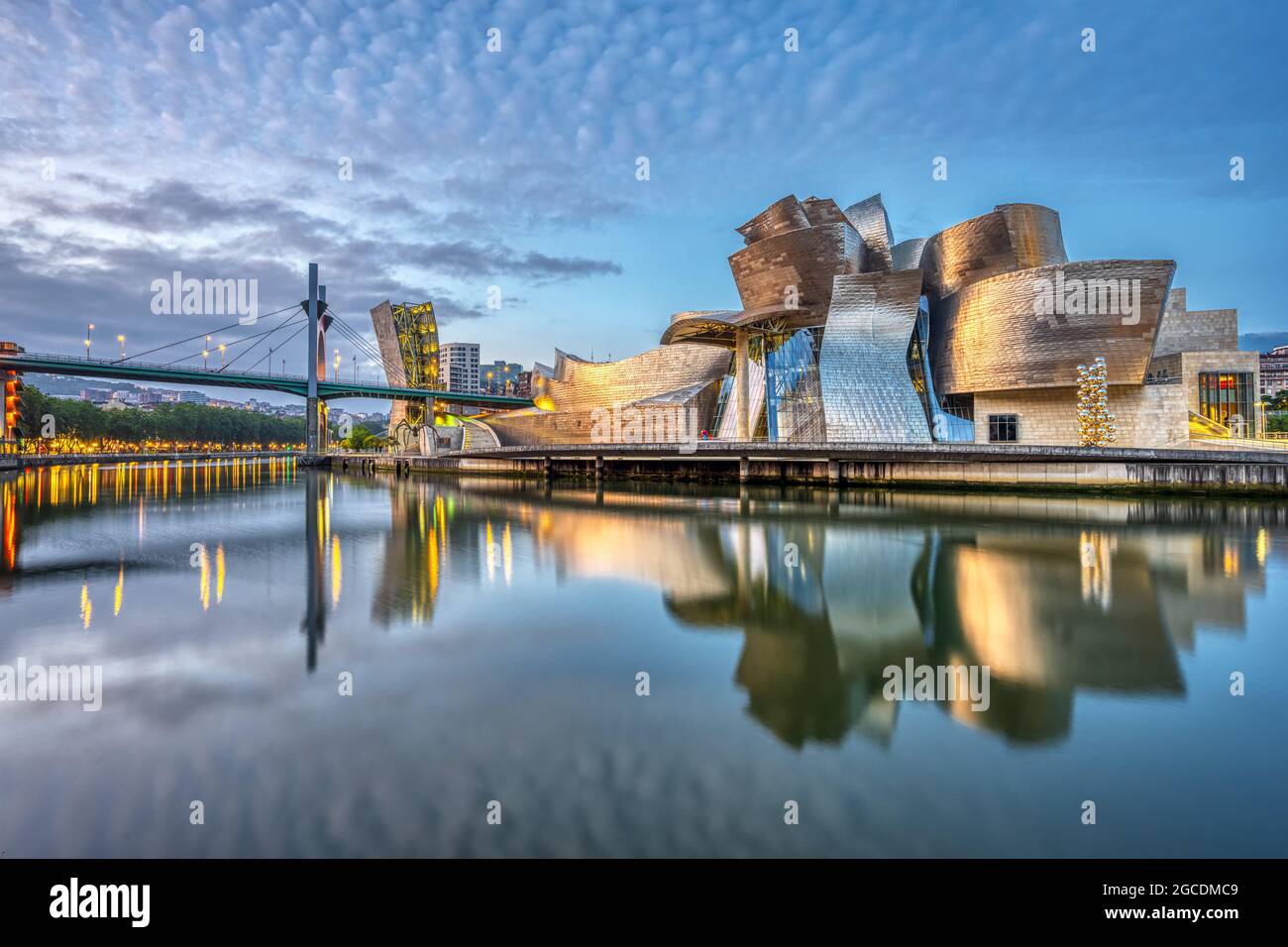 The Guggenheim Museum Bilbao and La Salve Bridge illuminated at dusk, reflected in the Nervión River, showcasing Bilbao's modern urban landscape.