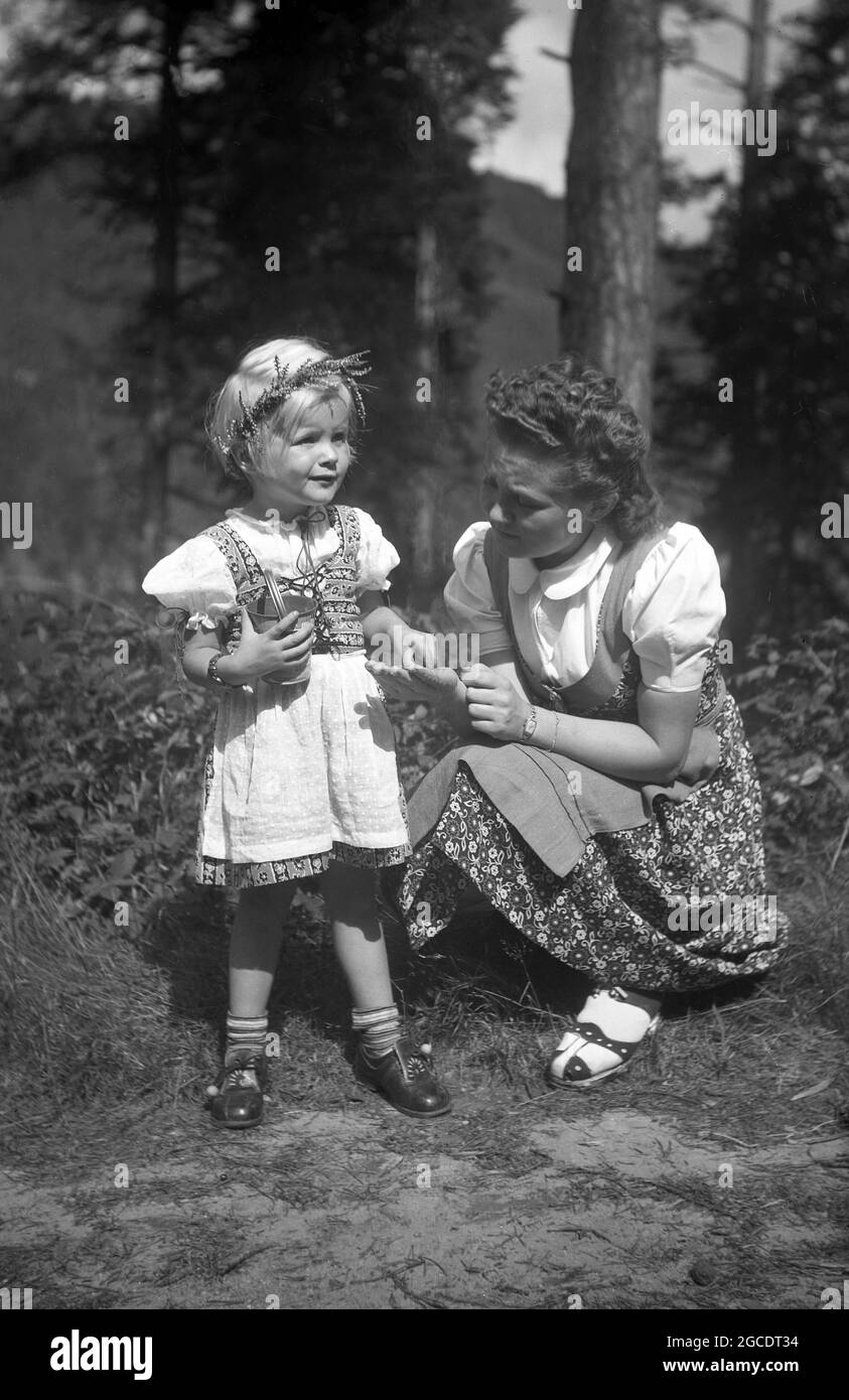 1950s, historical, a young girl with her mother picking wild flowers in a forest, Bavaria, Germany. Both are wearing a dirndl, a feminine dress, traditionally worn by women and girls in south-east Germany, Austria, Switzerland and other alpine areas. Regarded as a folk costume, it is derived from the clothing alpine peasants wore in the 16th century. To accompany the dirndl, a hair ornament, a smal floral wreath, known as a jungfernkranz is often worn and the young girl in the picture can be see to be wearing one. Stock Photo