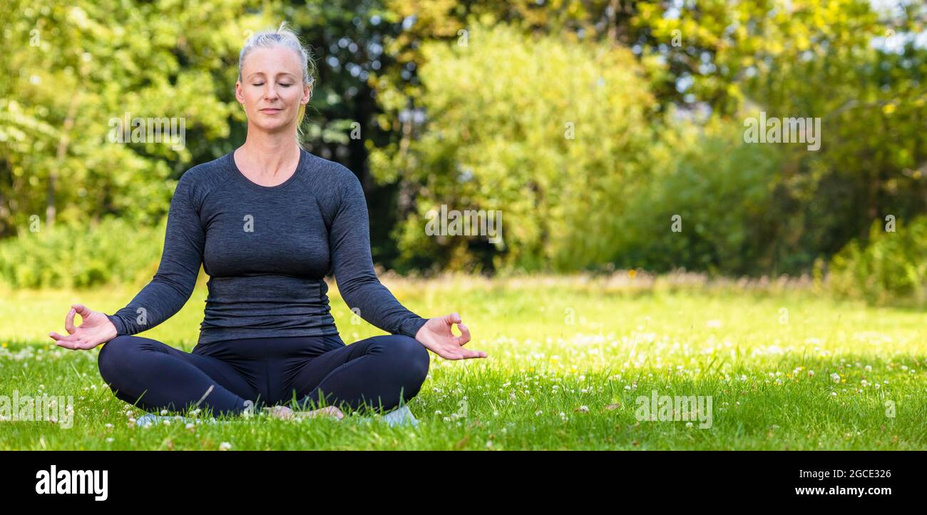 Panoramic web banner header of mature middle aged fit healthy woman practicing yoga outsidein a natural tranquil green environment panorama. Stock Photo