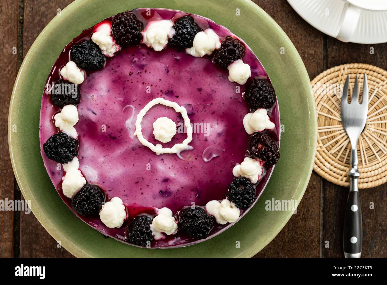 Top view of homemade blackberry cheesecake garnished with preserved blackberry and white cream in green ceramic plate on wooden table. Stock Photo
