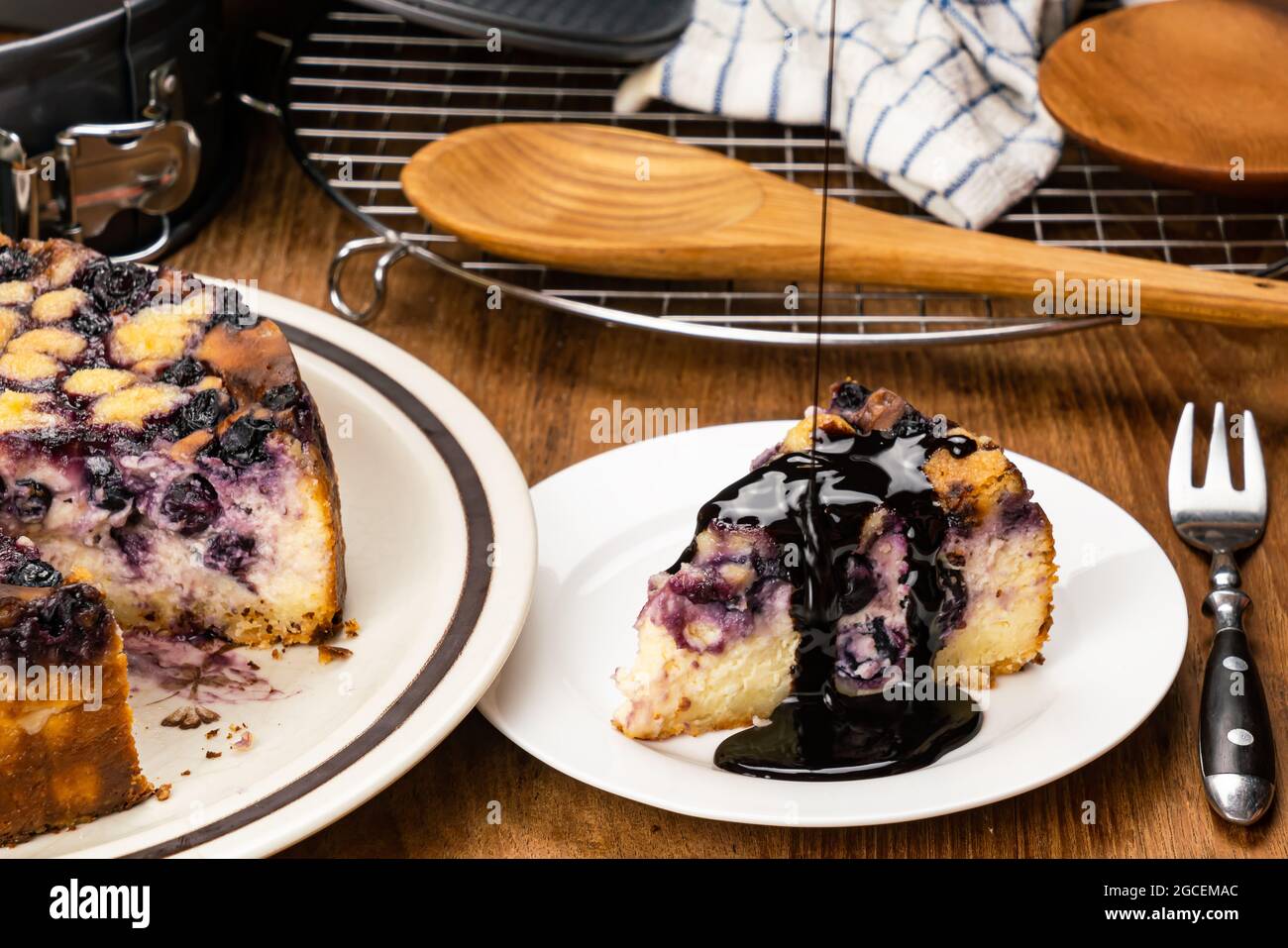 Pouring topping chocolate on a piece of delicious homemade blueberry and crumble cheesecake in white ceramic dish with a big one in brown dish, wooden Stock Photo