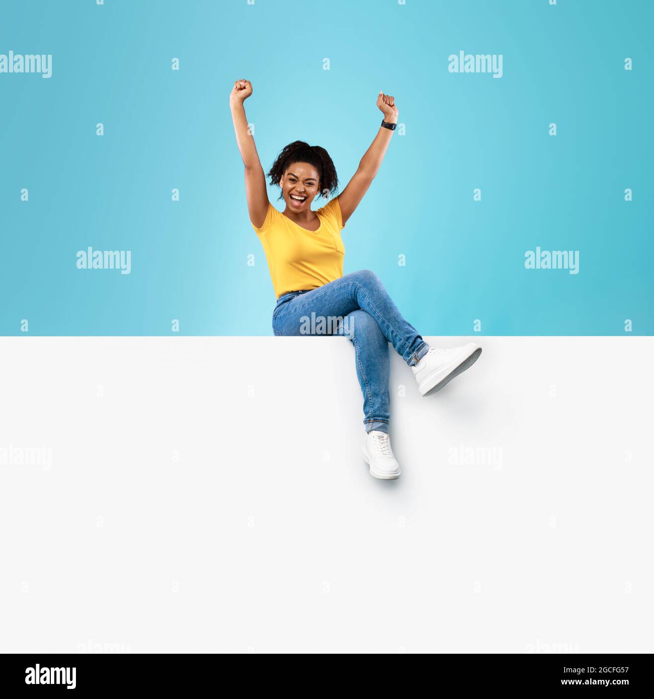 Portrait of excited young black lady sitting on blank white paper banner with mockup, raising hands up on blue studio background. Overjoyed woman with Stock Photo