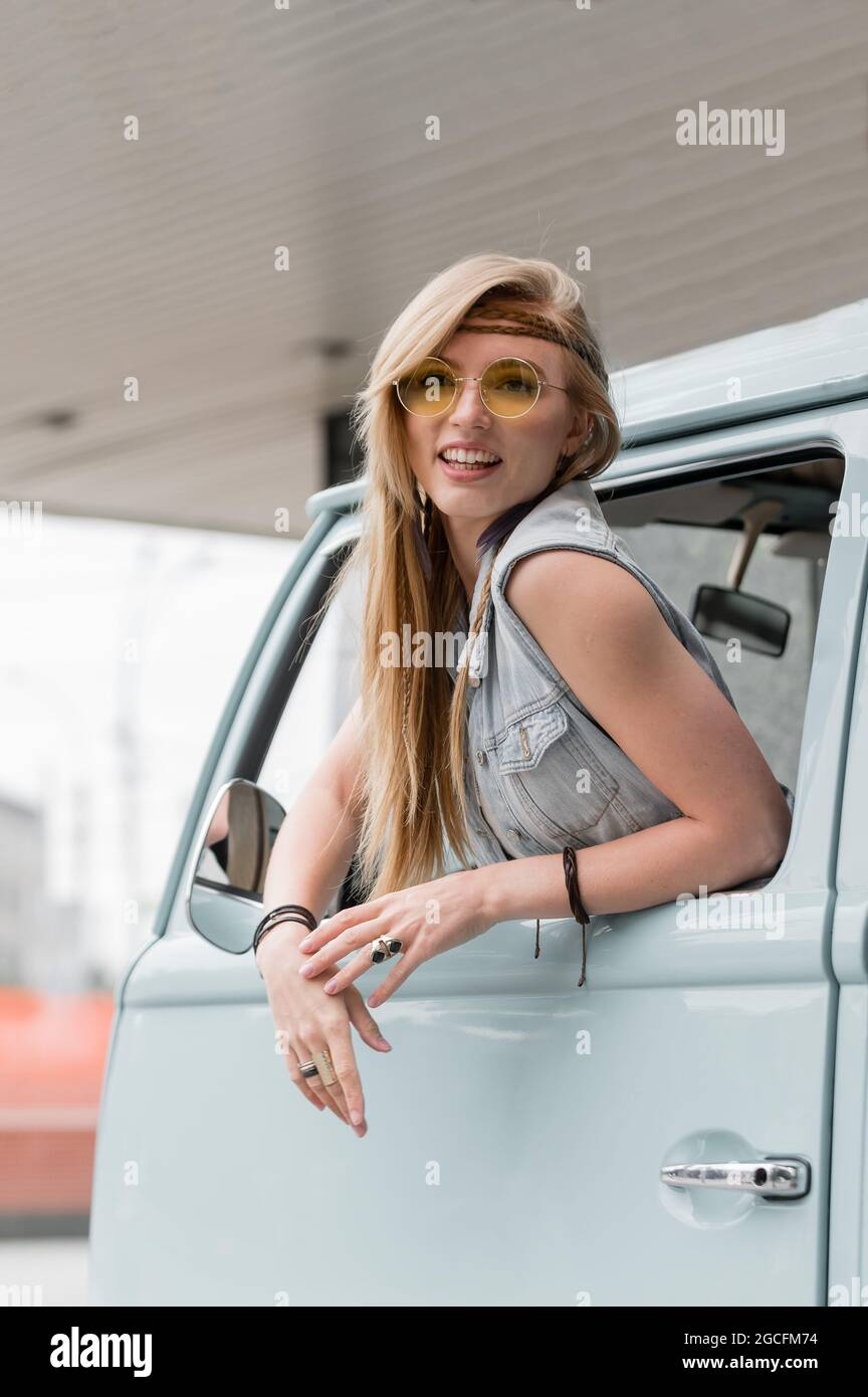 A beautiful young hippie woman in yellow sunglasses looks out the window of a blue vintage minivan. Make love, not war. Girl child flowers driving in Stock Photo