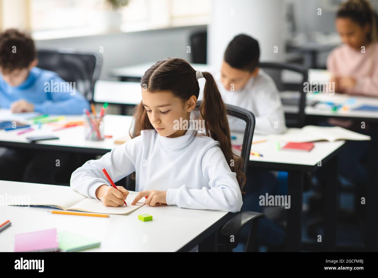 Cute female pupil sitting at desk in classroom at the elementary school. Student girl doing test in primary school. Children writing notes during the Stock Photo