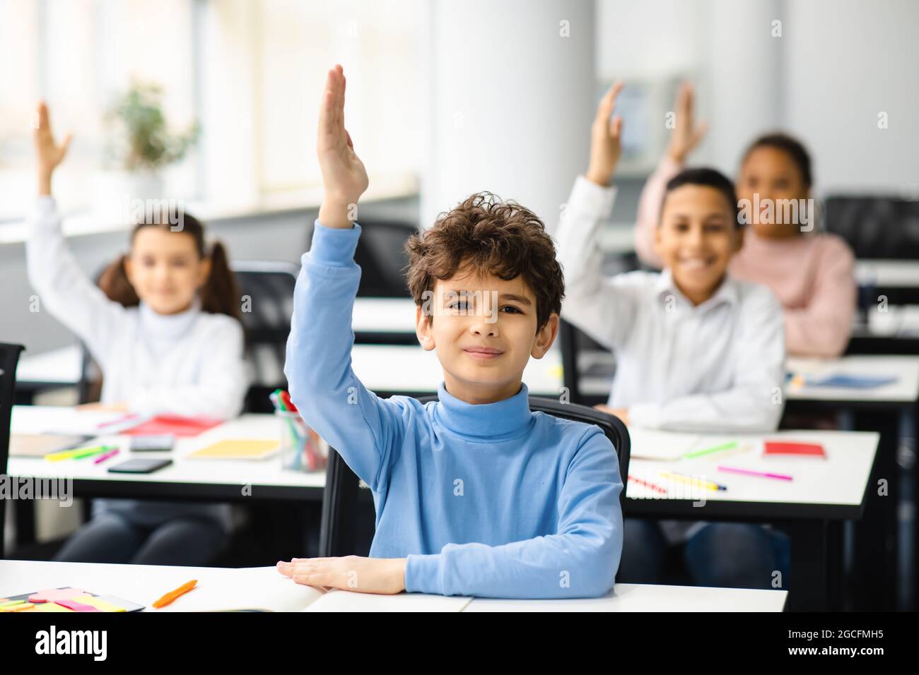 First Day Of School. Portrait of cute boy sitting at table in classroom, group of multicultural small pupils all raising hands up for an answer, study Stock Photo