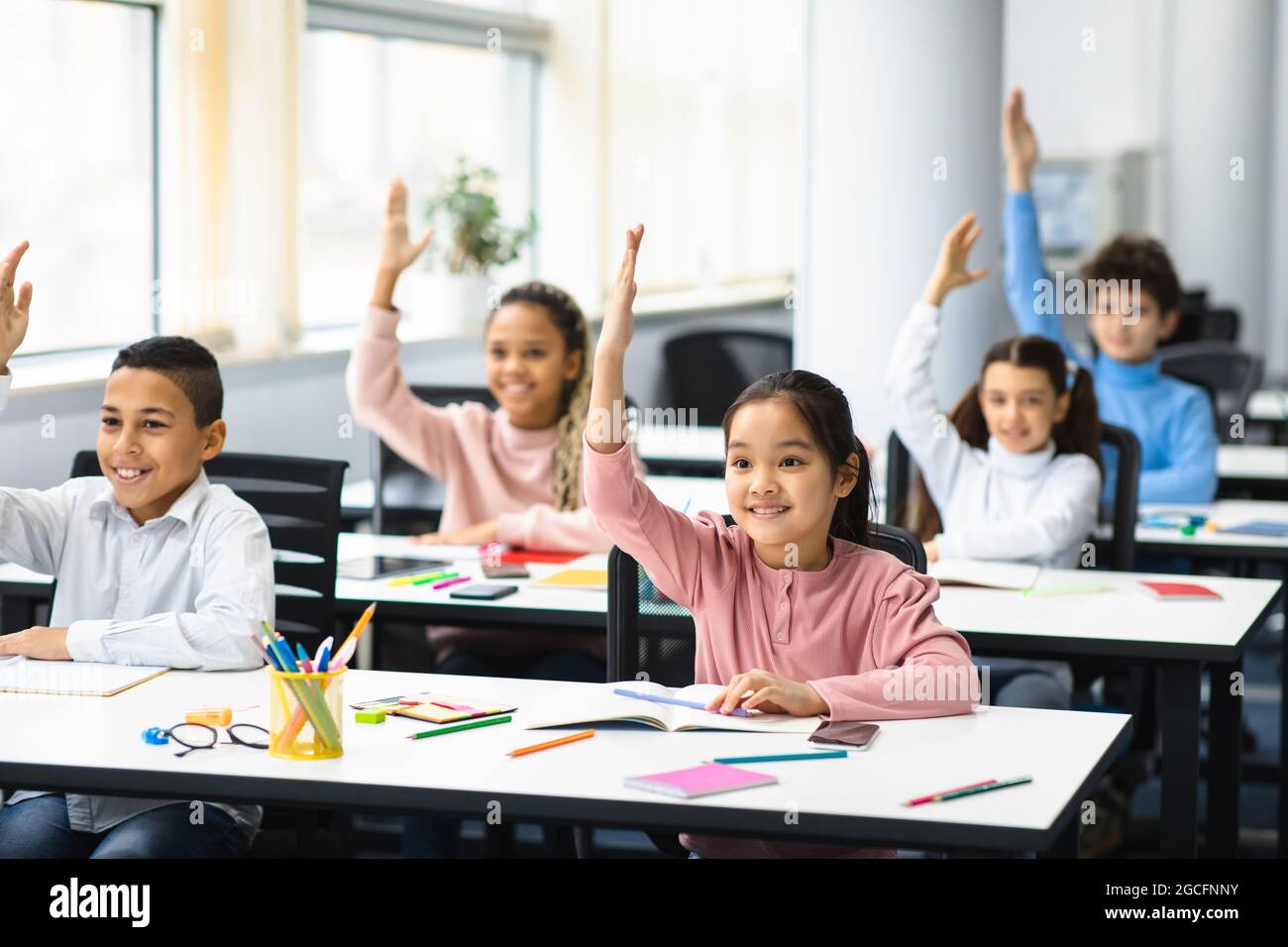 Return Back To School. Multiciltural diverse group of happy small elementary schoolchildren sitting at desks in classroom, raising hands up for an ans Stock Photo