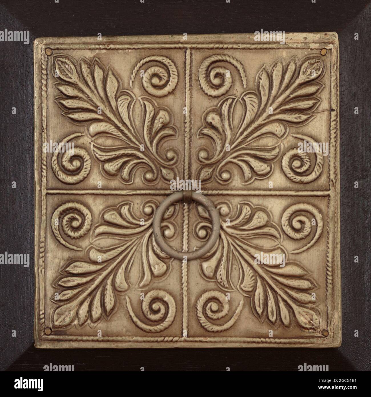 a square, decorative stamped metal panel of a dark wooden drawer with stylized botanical motifs and a circular pull in the center, in brown tones Stock Photo