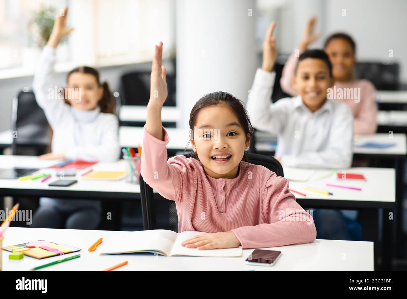 Concept of School Reopen, Back To Normal Lifestyle. Portrait of excited little schoolgirl raising hand up in the air to answer a question with all her Stock Photo