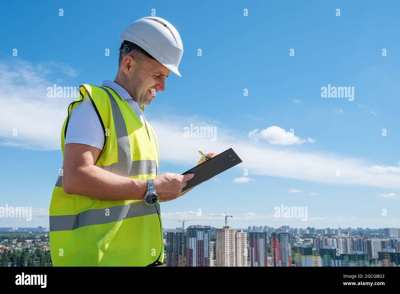 Smiling man in white helmet at construction site writes notes on clipboard Stock Photo
