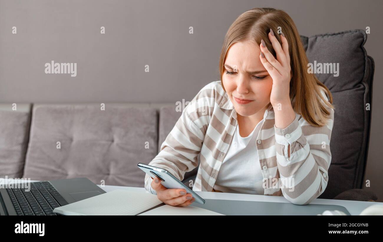 Young woman upset under stress. Confused woman holding head received bad news via smartphone at work. Teen girl looks at smartphone screen during Stock Photo