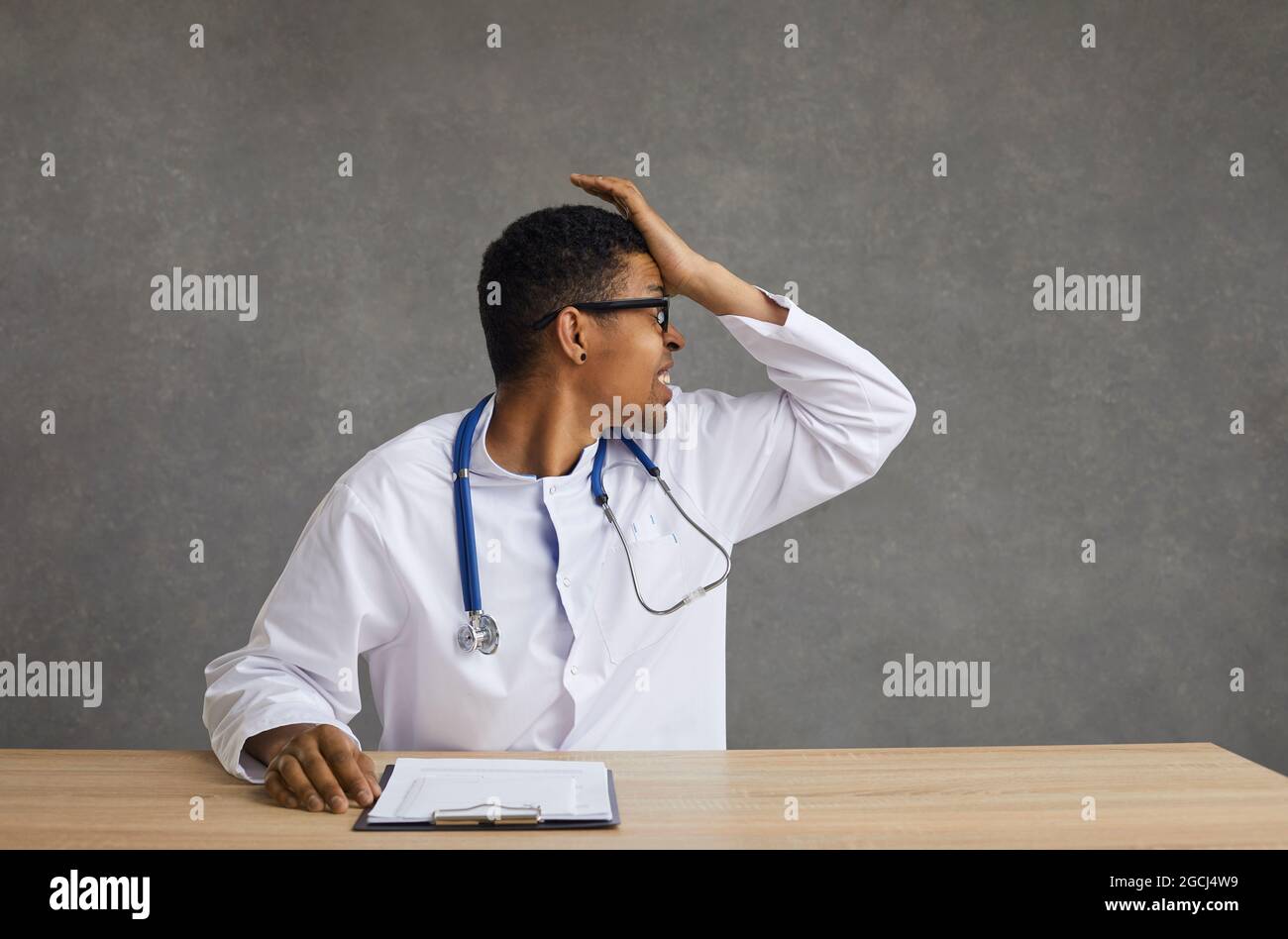 Worried frustrated doctor sitting at desk with hand on head studio shot Stock Photo