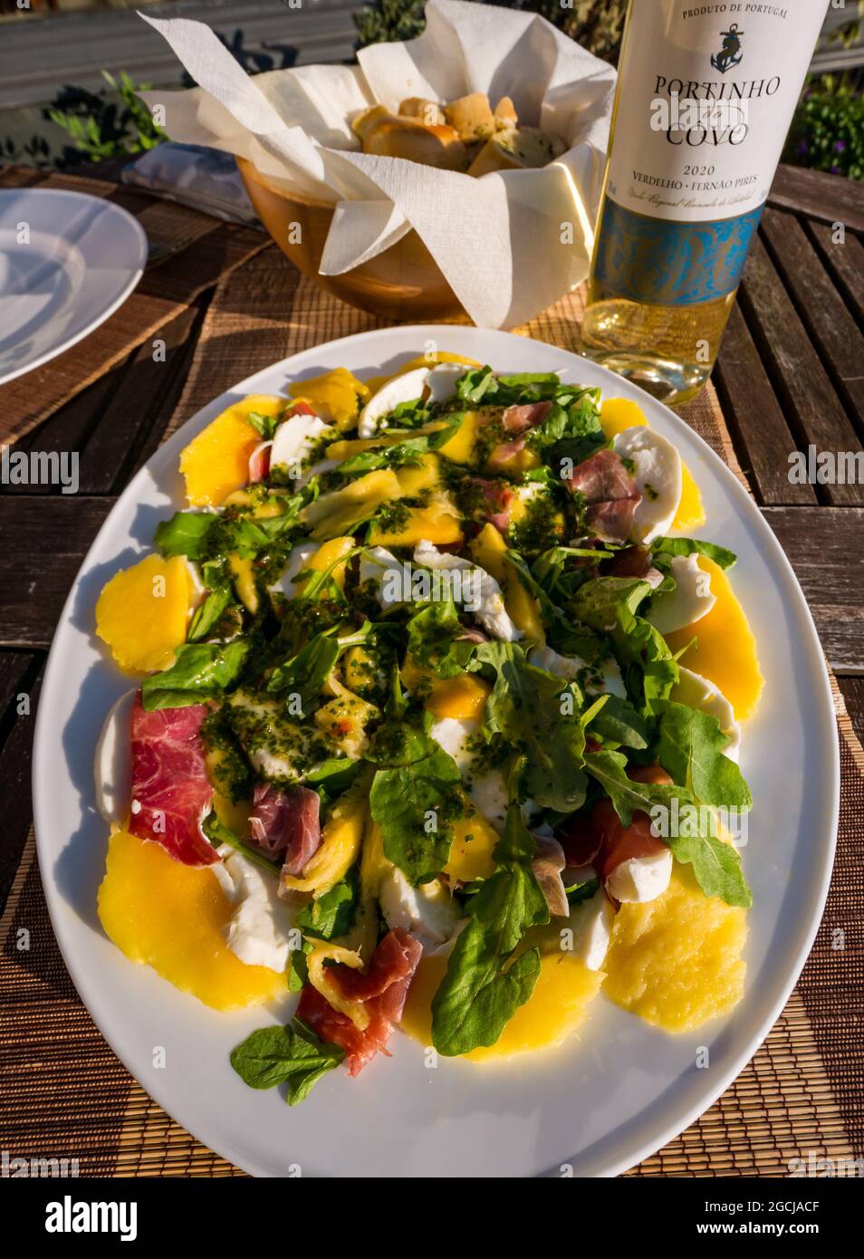 Summer salad platter with mango, prosciutto, basil & mozzarella cheese outdoors in sunshine with garlic bread and white wine bottle Stock Photo