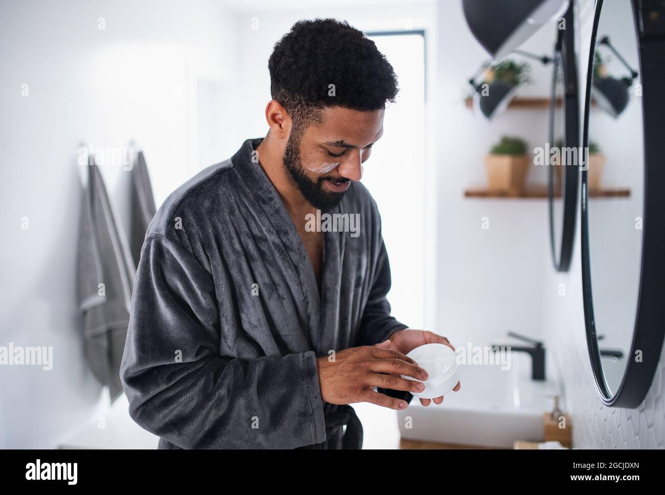 Young man applying cream on face indoors at home, morning or evening routine concept. Stock Photo