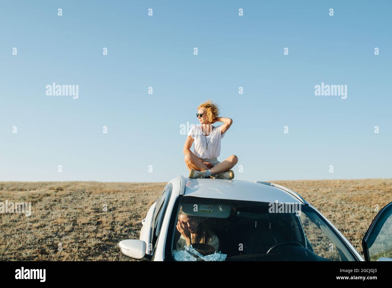 Joyous woman sitting on a car roof, enjoying wind and views on scenic grassy steppe plain. Her friend is in the car. Stock Photo