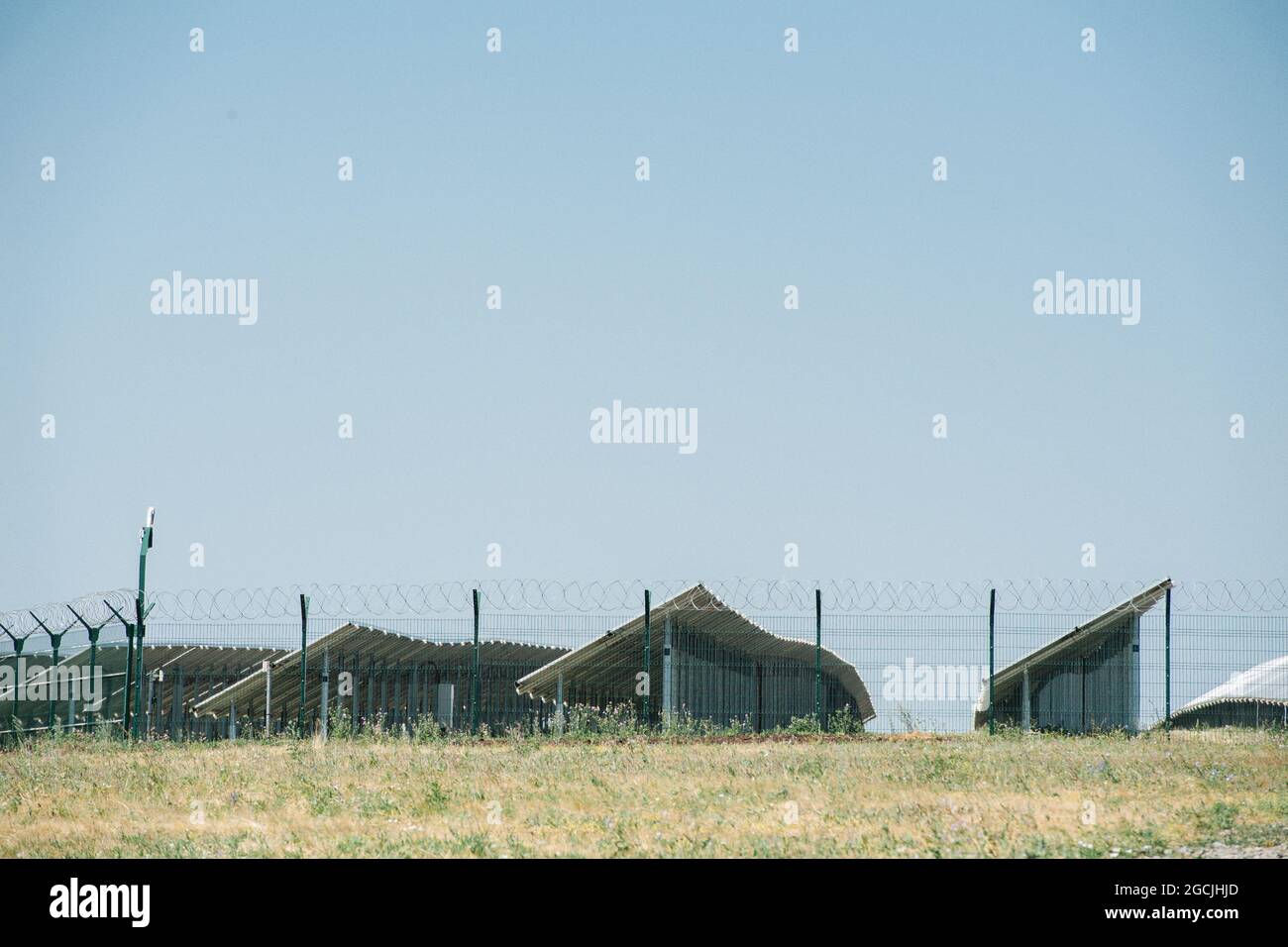 Multiple rows of solar panels behind a fence. Side view. Clear blue sky over them. Stock Photo
