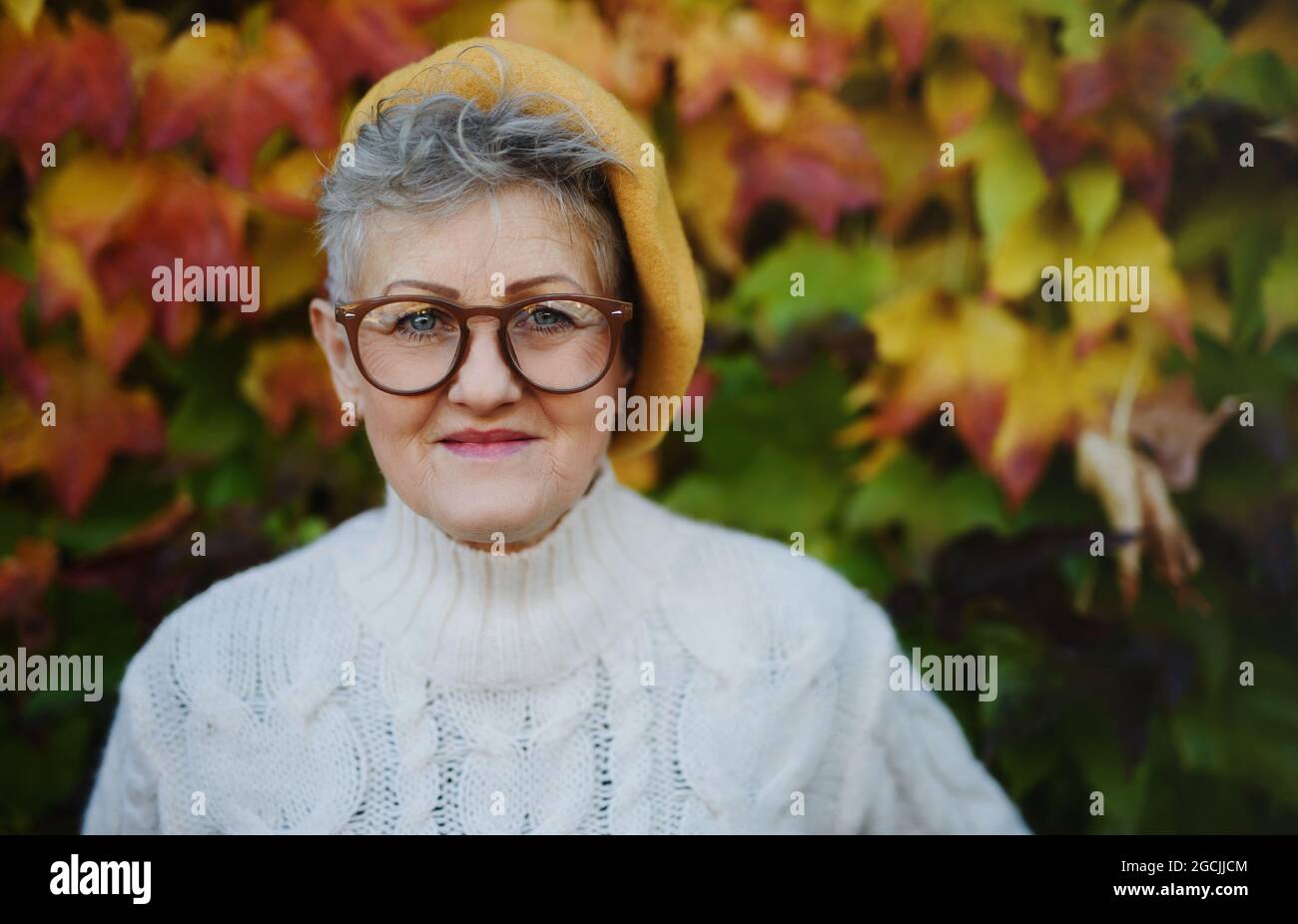 Senior woman standing outdoors against colorful natural autumn background, looking at camera. Stock Photo