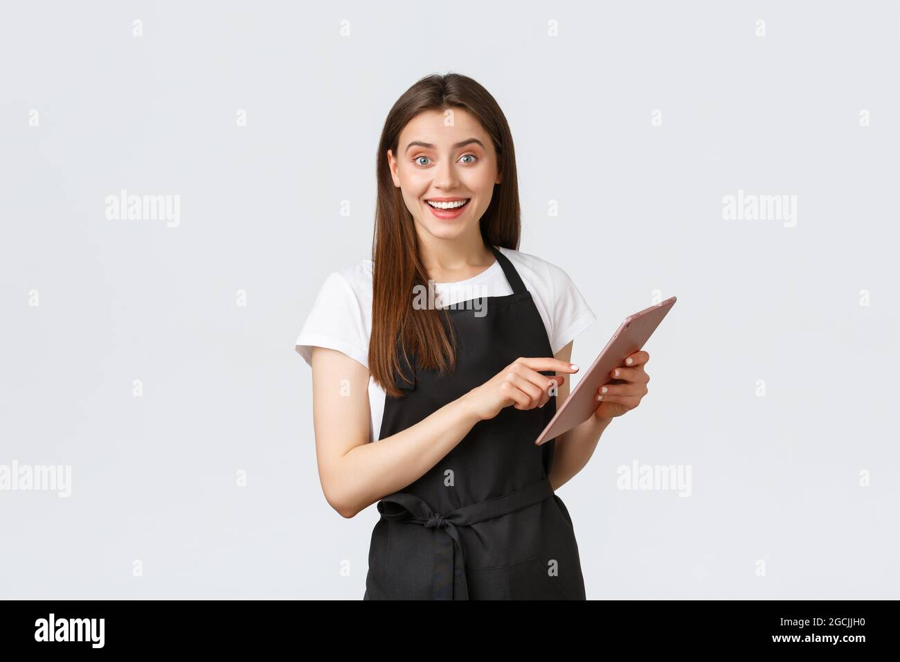 Grocery store employees, small business and coffee shops concept. Friendly cute female barista in black apron smiling at camera, using digital tablet Stock Photo