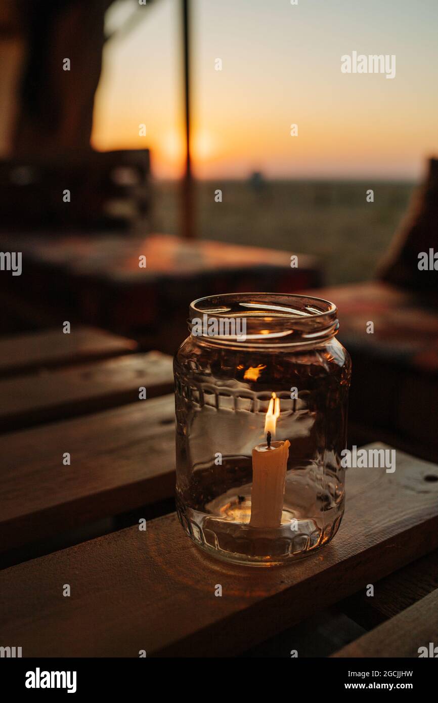 Candle burning inside a glass jar on a table. Sun setting over a steppe horison. Blurred background. Dark. Stock Photo