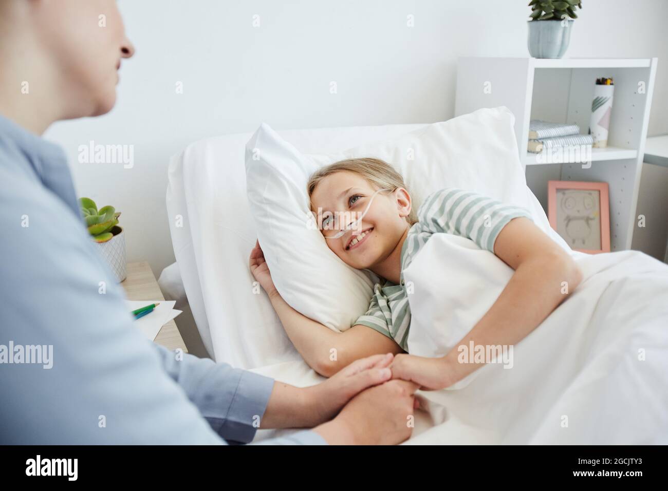 Portrait of little girl lying in hospital bed with oxygen support and smiling at mother caring for her Stock Photo
