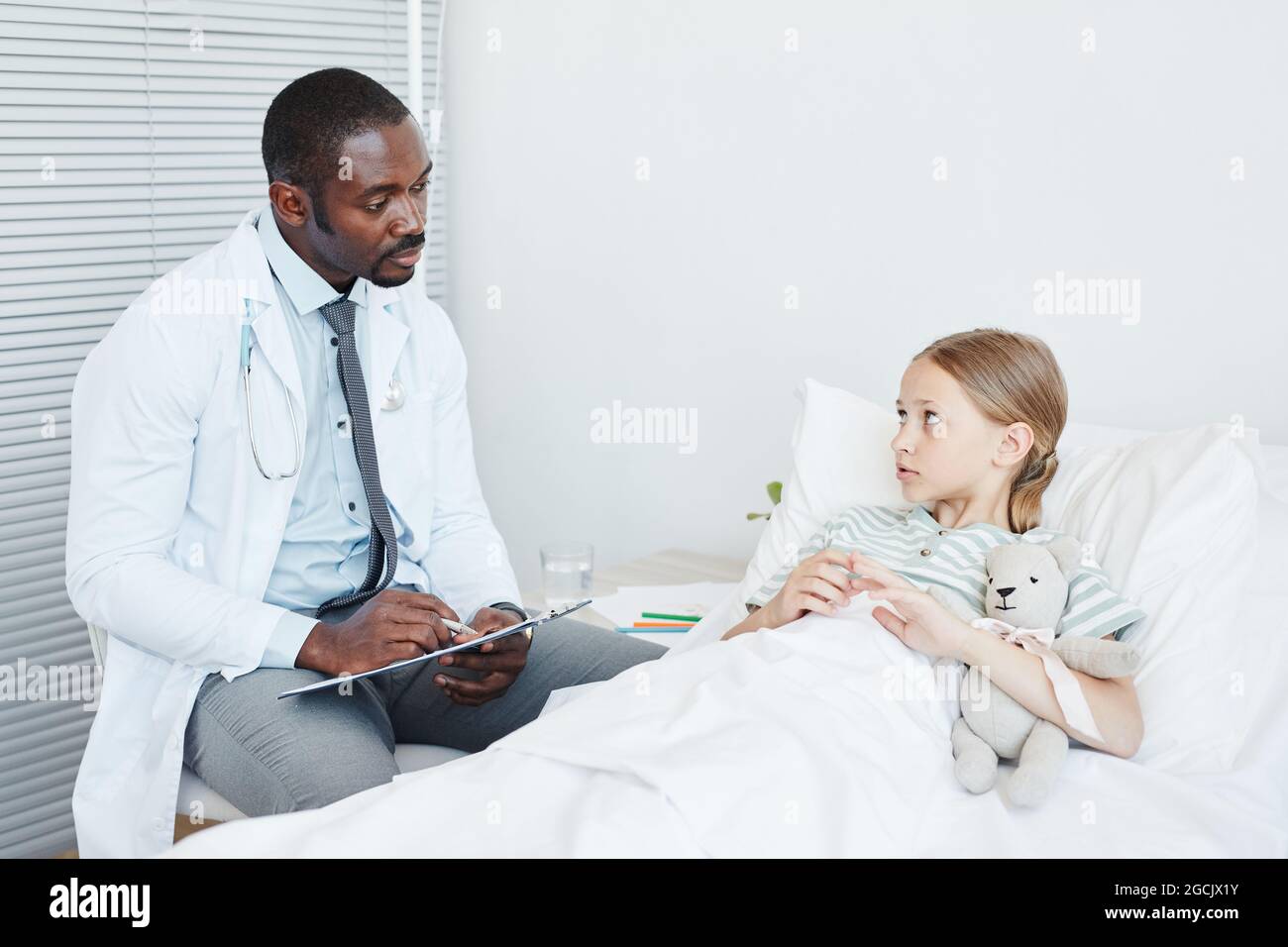 Portrait of young African-American doctor talking to little girl lying in hospital bed, copy space Stock Photo