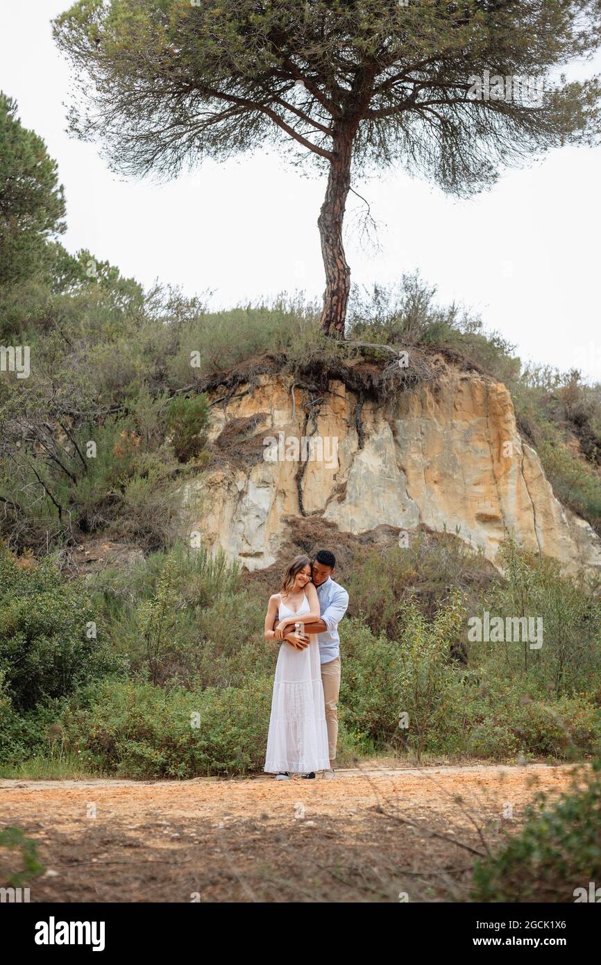 Happy diverse bride and groom embracing while standing in forest on wedding day Stock Photo