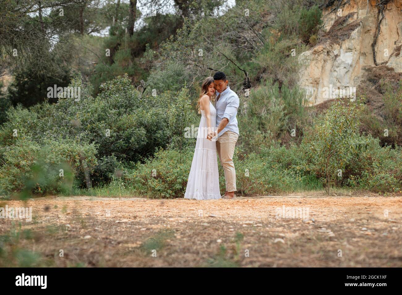 Diverse bride and groom embracing while standing in forest on wedding ...