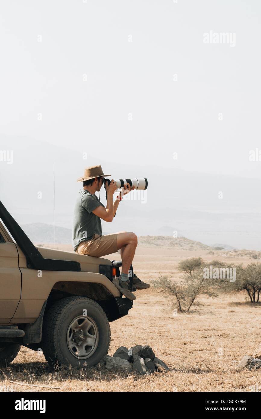 Side view of male traveling photographer sitting on offroader and taking picture on camera with telephoto lens during safari in savannah in summer Stock Photo