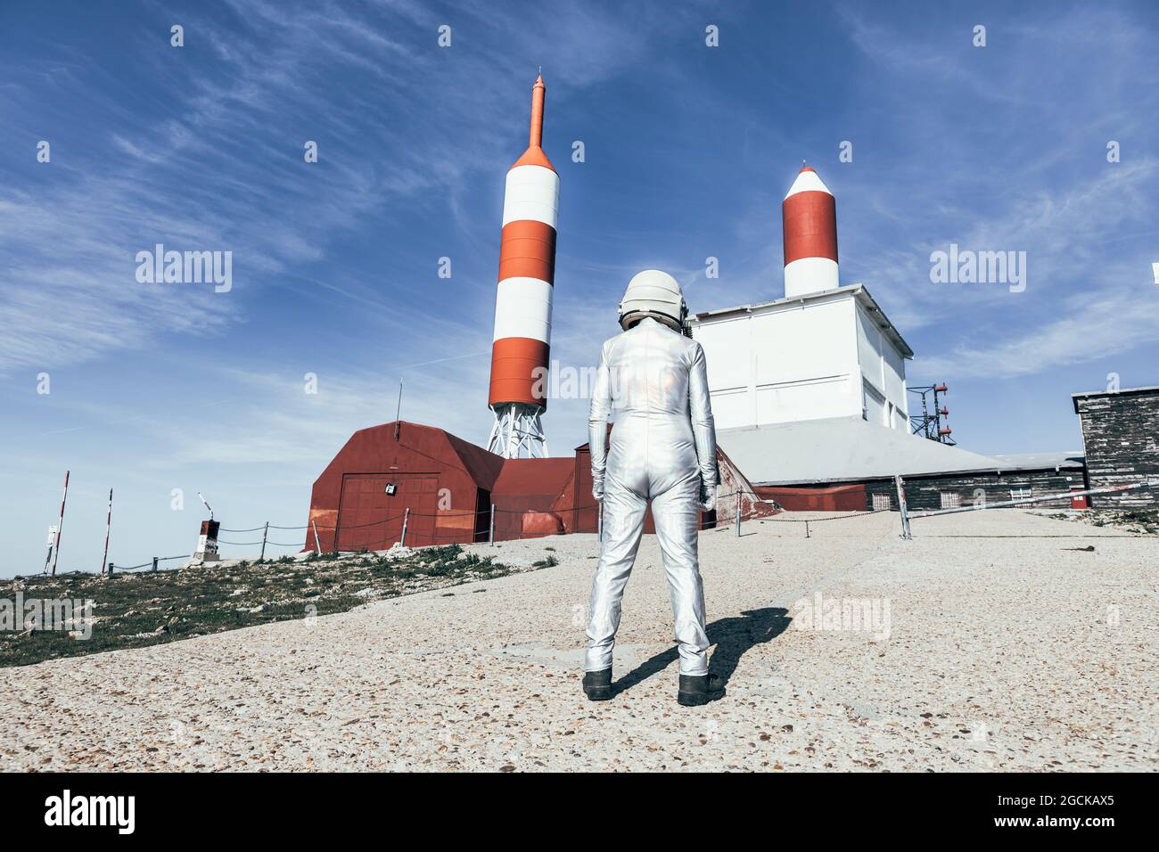 Back view man in spacesuit standing on rocky ground against metal fence and striped rocket shaped antennas on sunny day Stock Photo