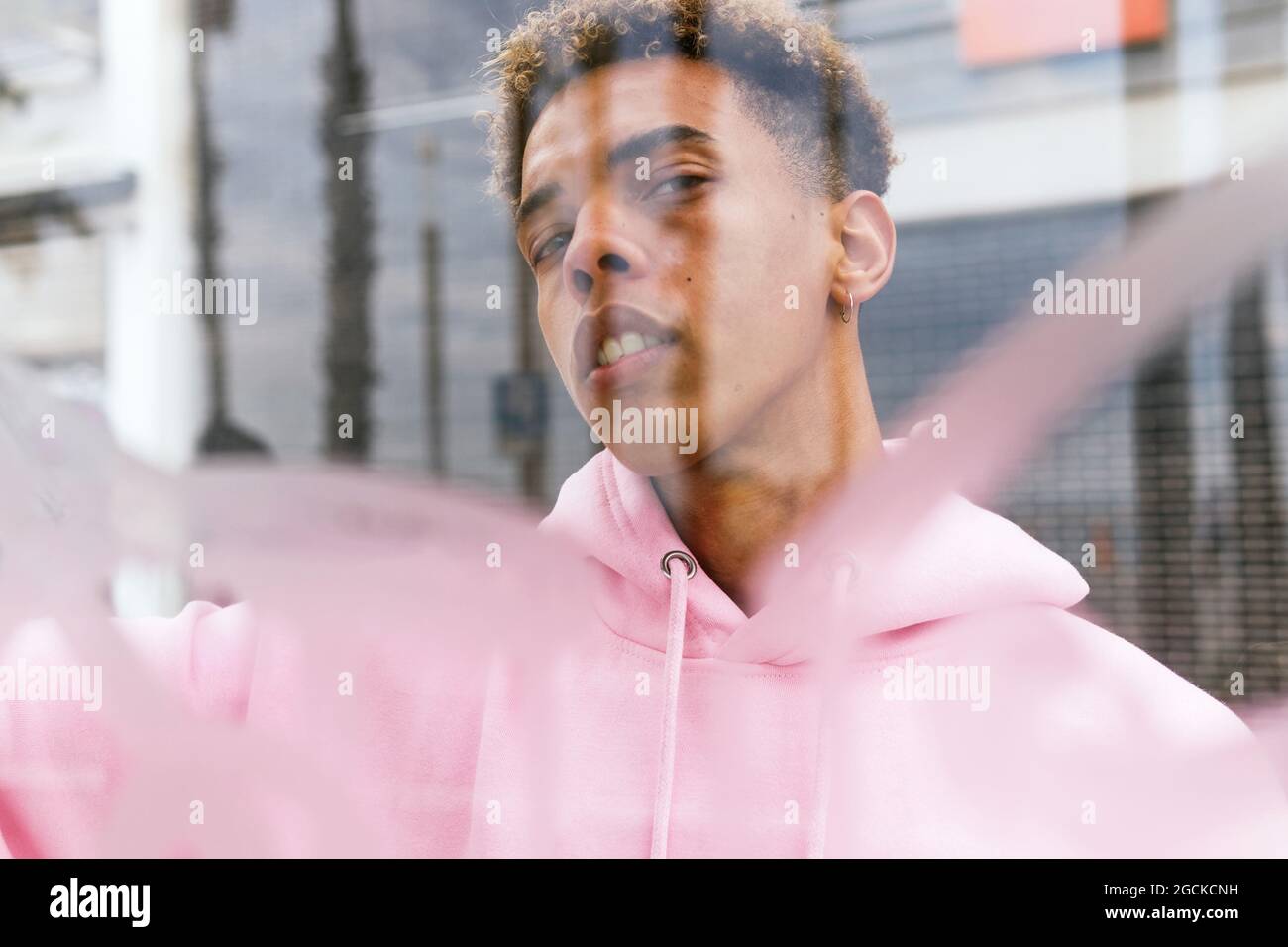 Through glass of serious young ethnic hipster guy with Afro hairstyle dressed in pink hoodie at window and looking at camera Stock Photo