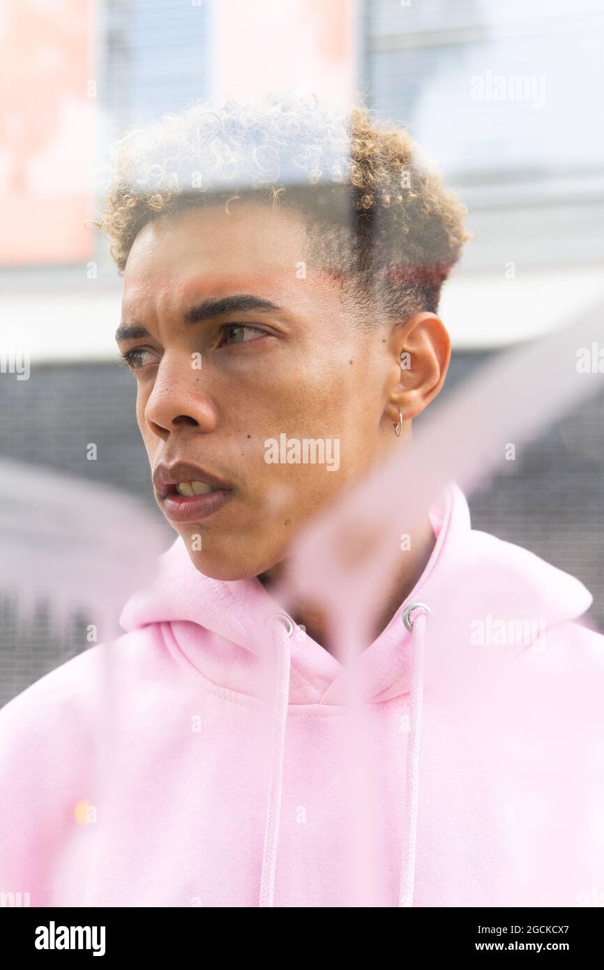Through glass of serious young ethnic hipster guy with Afro hairstyle dressed in pink hoodie at window and looking away Stock Photo
