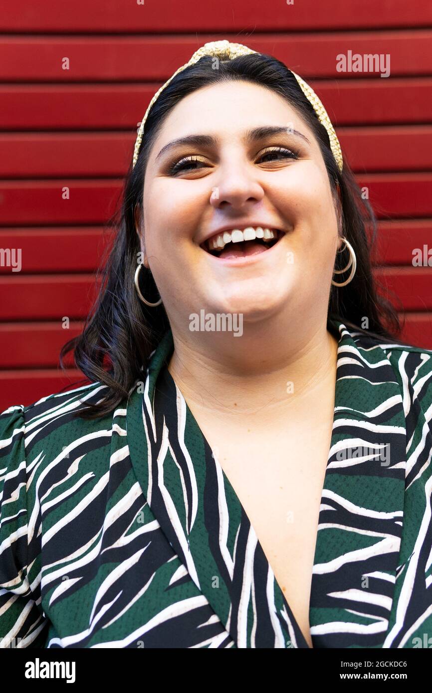Portrait of cheerful young curvy brunette in stylish striped outfit and  headband while looking at camera against red wall Stock Photo - Alamy